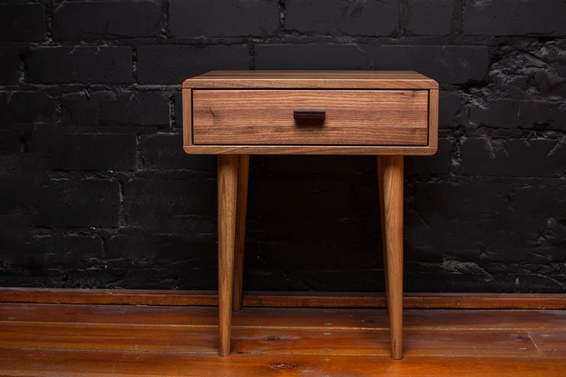 May include: A wooden nightstand with a single drawer and four legs. The drawer has a dark brown handle. The nightstand is made of light brown wood and has a natural finish.