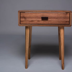 May include: A wooden nightstand with a single drawer and four legs. The drawer has a dark brown handle. The nightstand is made of a light brown wood.