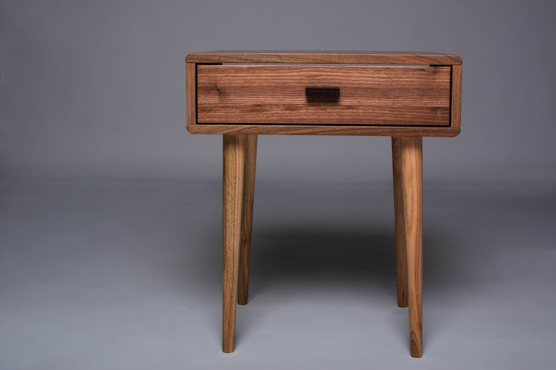 May include: A wooden nightstand with a single drawer and four legs. The drawer has a dark brown handle. The nightstand is made of a light brown wood.