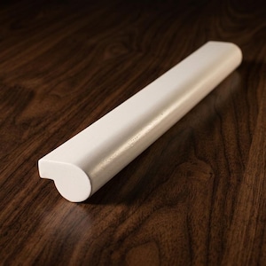 May include: A white, elongated wooden trim piece with a rounded end and a curved profile. The trim is set against a dark brown, wood-grain surface, creating a contrast in colour and texture. The design suggests a decorative or functional element.