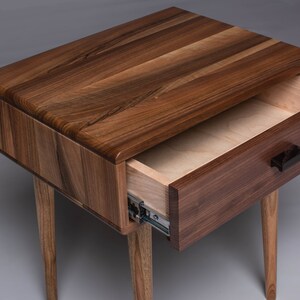 May include: A wooden nightstand with a single drawer that is open. The nightstand is made of walnut wood and has a dark brown finish. The drawer is made of maple wood and has a light brown finish. The drawer has a black wooden handle.