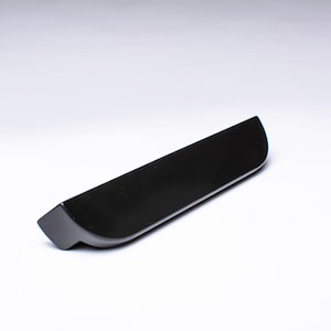 Può includere: Un oggetto nero rettangolare ed elegante con un bordo curvo. L'articolo ha una finitura opaca e un'estremità leggermente angolata. Sembra essere una maniglia o un elemento decorativo, forse per mobili o un'applicazione simile.