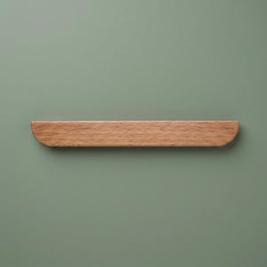 May include: A horizontal wooden cabinet handle with a light brown natural wood grain finish. The handle features a flat top edge and a rounded bottom edge, creating a semi-cylindrical shape that is smooth and uniform in its overall design.
