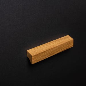 May include: A rectangular wooden cabinet pull with a light brown finish. The pull has a faceted design and a curved end, set against a dark background. This hardware is suitable for drawers and cabinets.