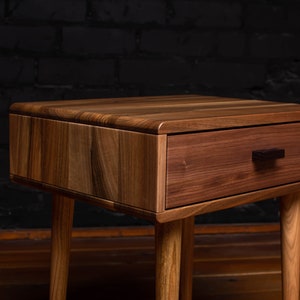 May include: A wooden nightstand with a single drawer and a dark wood finish. The drawer has a black handle. The nightstand has four legs.