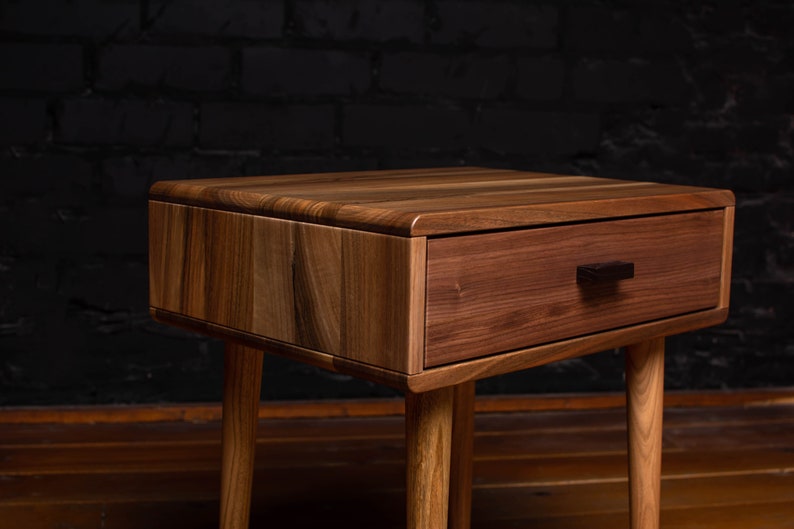 May include: A wooden nightstand with a single drawer and a dark wood finish. The drawer has a black handle. The nightstand has four legs.