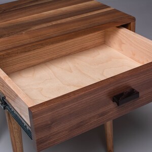 May include: A wooden nightstand with a single drawer that is open, revealing a light wood interior. The drawer has a black handle and the nightstand has four legs.