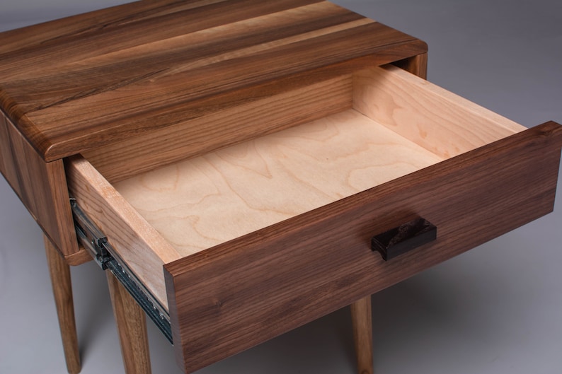 May include: A wooden nightstand with a single drawer that is open, revealing a light wood interior. The drawer has a black handle and the nightstand has four legs.
