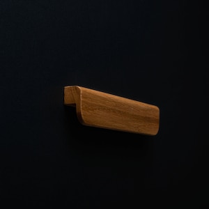 May include: A wooden drawer pull with a rounded rectangular shape. The pull is made of light brown wood and has a smooth finish.