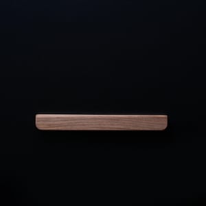 Peut inclure: Une poignée de tiroir en bois de forme rectangulaire arrondie. La poignée est de couleur brun clair et est placée sur un fond noir.