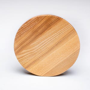 Pode incluir: Tábua de corte redonda de madeira clara, com veios visíveis. A tábua é castanho-clara e tem cerca de 30 cm de diâmetro. Está sobre um fundo branco.