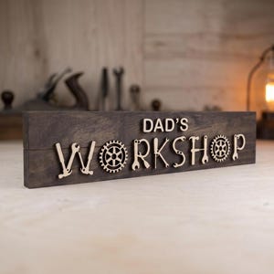 May include: A rectangular dark brown wooden sign featuring the text DAD'S WORKSHOP in light-colored wooden letters. The letters are creatively designed to incorporate shapes of tools and gears, including hammers, wrenches, and circular gear motifs.