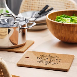 May include: Two cork trivets, one with a decorative design and the text "YOUR TEXT" printed on it. The trivets are on a light wood table with a stainless steel pot and a wooden salad bowl in the background.