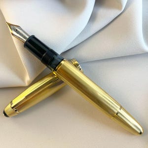 Könnte beinhalten: Ein goldener Füllfederhalter mit einem strukturierten Muster auf Schaft und Kappe. Der Stift hat einen schwarzen Griffbereich und eine silberne Feder. Die Kappe ist offen und gibt die Feder frei. Der Stift liegt auf einer weißen Oberfläche.