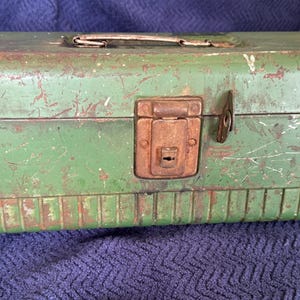 May include: A rectangular green metal toolbox featuring a weathered finish with visible scratches and rust. The box includes a central locking latch, two side latches, and a top handle. The lower section has a series of vertical embossed lines.