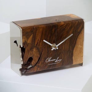 Puede incluir: Un reloj rectangular de madera con un acabado de veta natural. El reloj presenta un marco de metal plateado en los laterales y una esfera con manecillas plateadas. El texto "Chronos design" está inscrito en la madera.