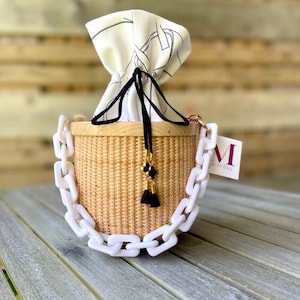 May include: A cylindrical woven basket bag with a light wood rim and a thick white acrylic chain handle. The bag features a white fabric drawstring closure with black line patterns and black tassels. A white tag with the letter M is attached.