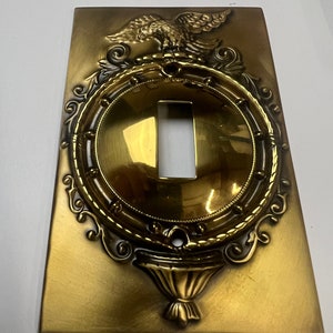 以下が含まれることがあります： 装飾的なワシ、ロープ状の縁取り、下部に装飾的な壺を備えた、金色のライトスイッチカバーです。