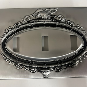 Puede incluir: Una placa de interruptor de metal plateado con un diseño ornamentado que presenta un águila y un marco ovalado negro. La placa tiene tres recortes rectangulares para interruptores.
