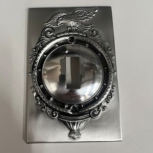 Puede incluir: Una cubierta de interruptor de luz de metal plateado con un diseño ornamentado que presenta un águila, un marco circular y una base decorativa.