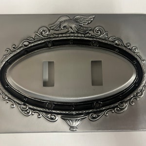 Puede incluir: Una cubierta de interruptor de luz decorativa plateada con un marco ovalado adornado y un diseño de águila en la parte superior.