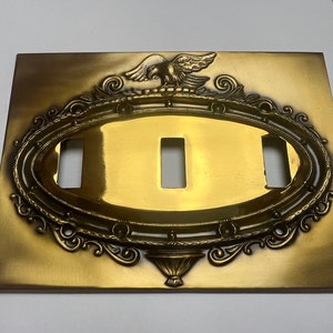 Puede incluir: Una tapa de interruptor de luz de metal dorado con un diseño ornamentado que presenta un águila y un marco ovalado decorativo.
