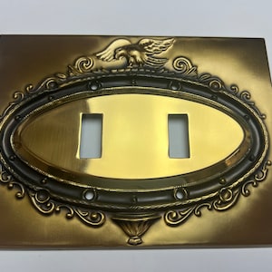 Puede incluir: Cubierta de interruptor de luz doble de latón dorado con un centro ovalado y borde decorativo. La cubierta presenta un águila en la parte superior y un jarrón en la parte inferior. Dimensiones aproximadas: 12 cm x 18 cm.