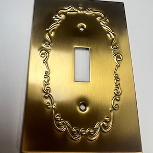 Könnte beinhalten: Ein goldfarbener Lichtschalter mit einem verzierten, geprägten Design. Die Abdeckung hat eine einzelne Schalteröffnung und zwei Schraubenlöcher.