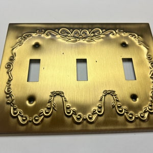 以下が含まれることがあります： 真鍮色の装飾的なトリプルライトスイッチプレート。プレートは長方形で、スイッチ用の長方形の切り欠きが3つあります。縁には、華やかなエンボス加工の花柄のデザインが施されています。