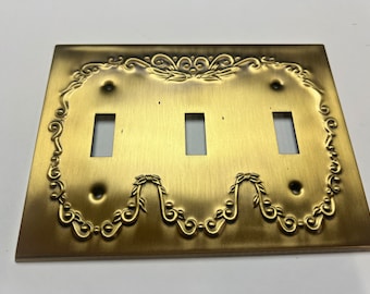 May include: A decorative brass-colored triple light switch plate. The plate has a rectangular shape with three rectangular cutouts for the switches. It features an ornate, embossed floral design around the edges.