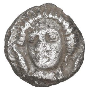 Puede incluir: Una moneda de plata con un relieve detallado de un rostro humano, cabello rizado y ojos grandes. La moneda tiene una apariencia desgastada y antigua, con imperfecciones visibles y una forma irregular. El rostro está centrado en la moneda.
