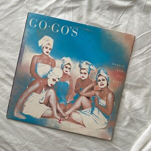 May include: Vintage vinyl record album cover for "Beauty and the Beat" by The Go-Go's. The cover features a group of women in white towels and head wraps, with blue and pink accents. The album title is in large white letters.