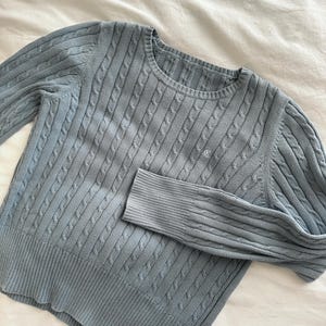 May include: A light blue cable knit sweater with a crew neck and long sleeves. The sweater features a small embroidered detail on the chest. The fabric has a textured, ribbed pattern. The sweater is made of a soft, comfortable material.