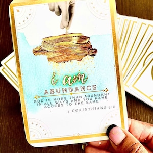 Pode incluir: Um cartão de oráculo com bordas douradas com as palavras "i am ABUNDANCE". A imagem mostra uma mão derramando pó de ouro sobre uma pilha de ouro. O texto "GOD IS MORE THAN ABUNDANT" também está presente. Parte de um baralho de cartas de oráculo.