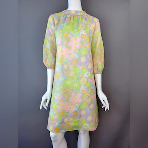 May include: A vintage floral print dress in pastel colors. The dress features a high neckline, three-quarter sleeves, and a knee-length hem. The fabric is a sheer material with a pattern of pink, yellow, green, and purple flowers. The dress is displayed on a mannequin.