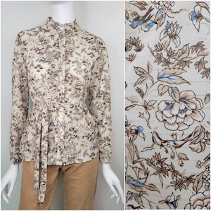 May include: A beige long-sleeved blouse with a floral print featuring brown and blue flowers, birds, and gold accents. The blouse has a high collar and a tie at the waist.