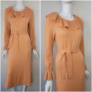 May include: A peach-colored knitted dress with a ruffled collar and cuffs. The dress has a self-tie belt at the waist.
