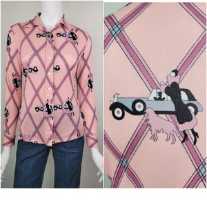 May include: A pink long-sleeved button-up shirt with a plaid pattern and a repeating design of a car with a couple inside and two dogs. The shirt has a collar and long sleeves.