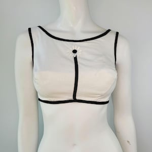 May include: A white sleeveless top with black trim and a daisy accent. The top features a sheer overlay and a black vertical stripe with a daisy applique. The neckline and armholes are outlined in black.