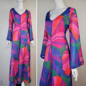 Puede incluir: Un vestido largo y fluido con cuello en V y mangas largas acampanadas. El vestido presenta un vibrante estampado abstracto en tonos de rosa, morado, naranja, verde y azul. La tela parece ligera y tiene un brillo sedoso, creando una estética retro y bohemia.