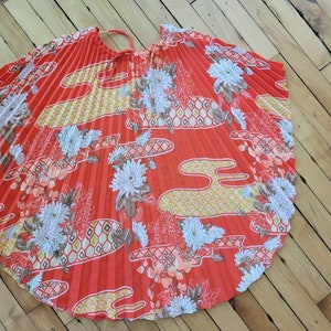 以下が含まれることがあります： 鮮やかな赤色のプリーツ加工が施された円形の衣服。白、黄色、桃色の花柄と雲模様で装飾されています。上部には細いストラップが付いています。このデザインは、ヴィンテージ感を醸し出しています。