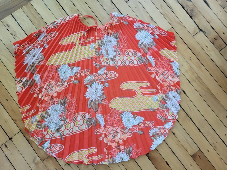 以下が含まれることがあります： 鮮やかな赤色のプリーツ加工が施された円形の衣服。白、黄色、桃色の花柄と雲模様で装飾されています。上部には細いストラップが付いています。このデザインは、ヴィンテージ感を醸し出しています。