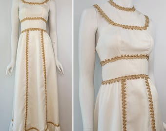 Puede incluir: Un vestido blanco sin mangas, largo hasta el suelo, con adornos dorados. El vestido presenta adornos dorados alrededor del cuello, la cintura y en la parte delantera. El dobladillo tiene un detalle de volantes. El vestido se exhibe en un maniquí.