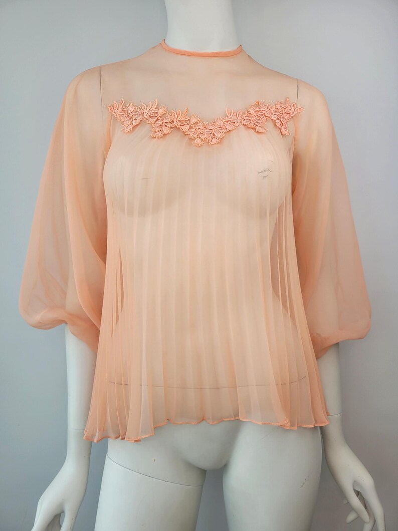 May include: Peach-colored sheer blouse with a high neckline and short, puffy sleeves. The blouse features a pleated front and a delicate floral lace detail across the chest. The fabric appears lightweight and flowing, creating a soft, feminine look.