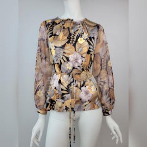 May include: A vintage floral print blouse with long, sheer sleeves. The blouse features a round neckline, a tie waist, and a button-down front. The print includes flowers and leaves in shades of gold, brown, black, and gray.