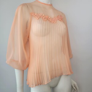 May include: A peach-colored, sheer blouse with a high neckline and three-quarter sleeves. The blouse features a pleated front and a delicate floral lace detail across the upper chest. The fabric appears lightweight and flowing.