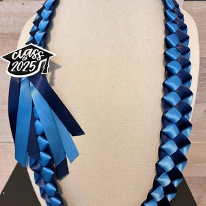 May include: A graduation lei in shades of blue, featuring woven ribbon and a decorative accent. The lei has a charm that reads "Class of 2025" in a graduation cap design, with matching ribbons.
