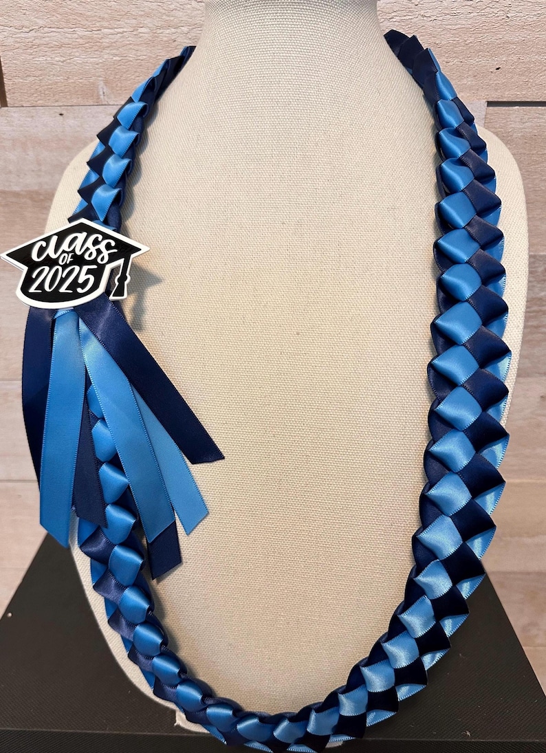 May include: A graduation lei in shades of blue, featuring woven ribbon and a decorative accent. The lei has a charm that reads "Class of 2025" in a graduation cap design, with matching ribbons.