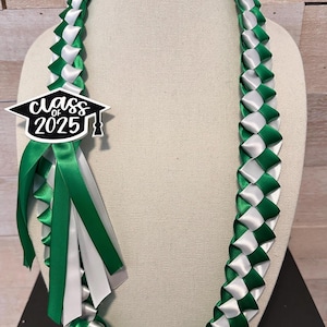 May include: A graduation lei in green and white ribbon, displayed on a mannequin. The lei features a woven pattern and a charm that reads "Class of 2025" in black and white. Green and white ribbons hang from the charm.