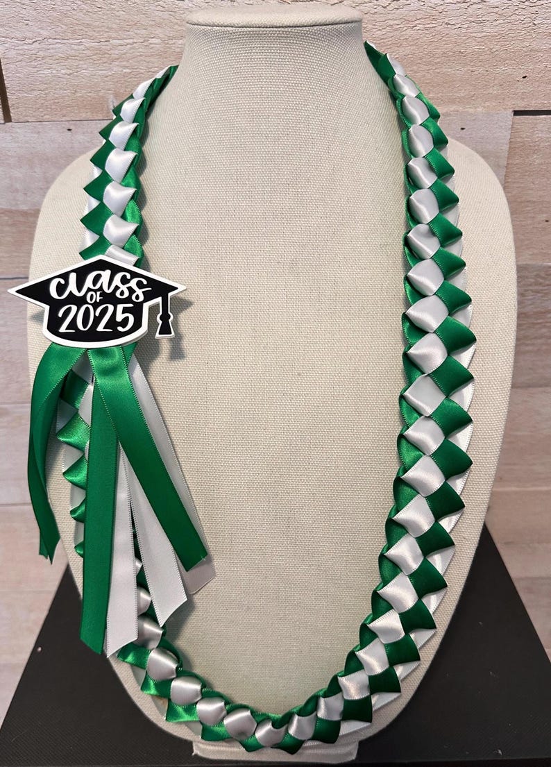 May include: A graduation lei in green and white ribbon, displayed on a mannequin. The lei features a woven pattern and a charm that reads "Class of 2025" in black and white. Green and white ribbons hang from the charm.